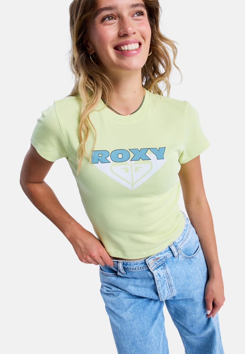 Jasnożółta dopasowana koszulka z niebieskim logo i grafiką "ROXY", zestawiona z jasnoniebieskimi dżinsami z wysokim stanem.