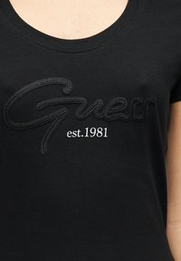 Μαύρο βαμβακερό μπλουζάκι με στρογγυλή λαιμόκοψη. Διαθέτει ανάγλυφο λογότυπο "Guess" και "est. 1981" σε λευκό με αντίθετο χρώμα. Λεπτή υφή.