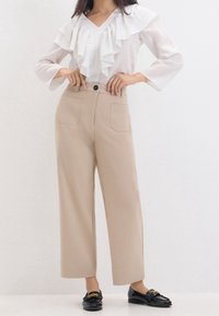 Kvinde iført beige brede bukser med forlommer, en hvid bluse med flæser og sorte loafers med gulddetaljer, stående med hænderne på hoften.