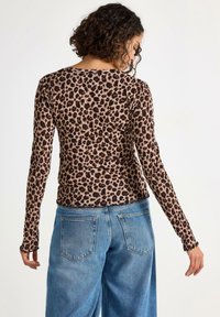 Žena s kudrnatými vlasy, oblečená v přiléhavém topu s leopardím potiskem s dlouhým rukávem a volných modrých džínách, otočená zády k jednobarevnému pozadí.