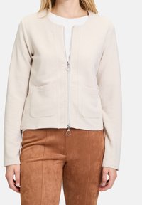 Light beige zip-up sweatshirt with a round neckline, front pockets, ribbed texture, and a silver zipper pull, paired with brown suede-like pants.