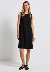 Robe noire sans manches à coupe droite, en tissu doux, arrivant au genou, avec un col rond, accompagnée de sandales tressées noires.