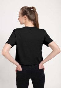 Černé tričko s krátkým rukávem vyrobené z bavlny. Jednoduchý design s kulatým výstřihem. Model stojí se založenýma rukama v kapsách černých kalhot.
