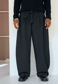 Sorte bukser med vide ben og bindebælte, der har en glat stoftstruktur og plisseret design. Kombineret med sorte sko.