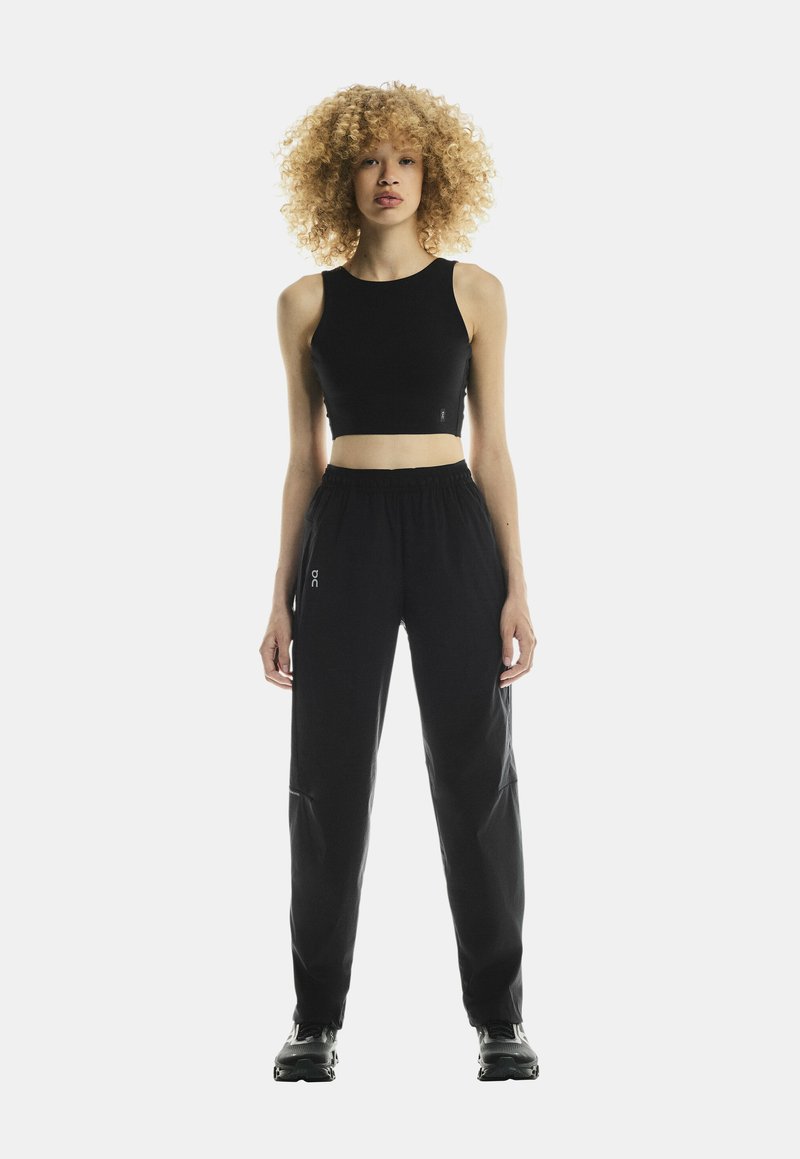 Black crop top with a round neckline, paired with black elastic-waist pants featuring pockets and a logo detail, worn with black sneakers.