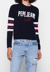 Pull en tricot bleu marine avec des manches rayées blanches et rouges. Il présente les inscriptions "PEPE JEANS" et "Since 1973" en texte contrastant. Associé à un jean bleu clair déchiré.