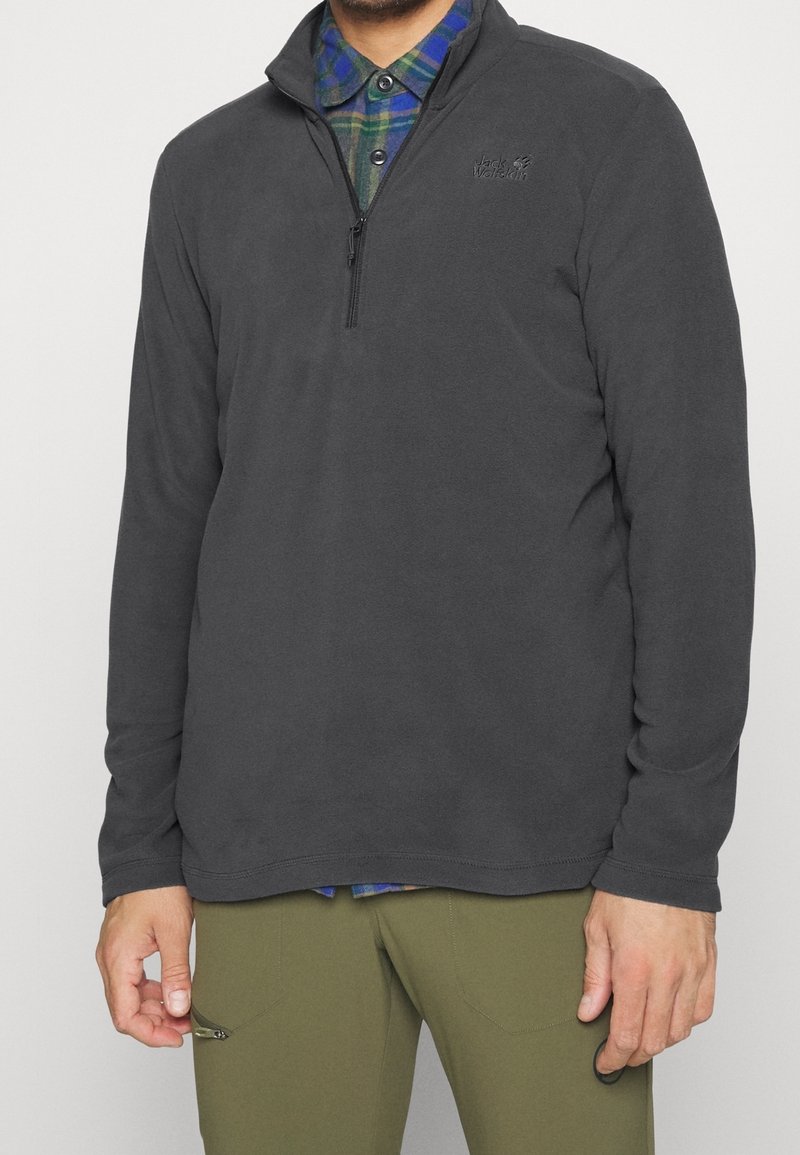Tummanharmaa fleece-neule, jossa on puolivetskari ja logoyksityiskohta, päällä sinivihreä ruudullinen paita, yhdistettynä oliivinvihreisiin housuihin.