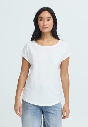 Vrouw met lang donker haar, gekleed in een effen wit T-shirt met korte mouwen en lichtblauwe spijkerbroek, staand tegen een lichte achtergrond.