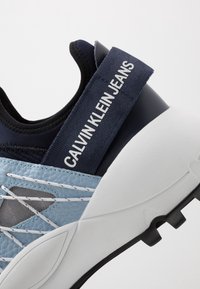Modré a námořnické tenisky s texturovaným svrškem, bílými tkaničkami a gumovou podrážkou. Obsahují značkový pásek s nápisem "CALVIN KLEIN JEANS".