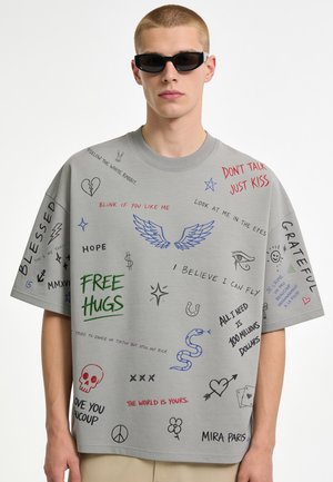 Junger Mann mit Sonnenbrille und grauem T-Shirt, bedeckt mit bunten handgeschriebenen Worten, Symbolen und Zeichnungen wie Flügeln, Herzen und einer Schlange.