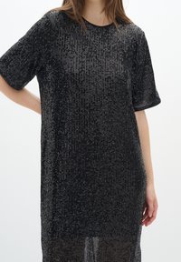 Svart paljettklänning med en avslappnad silhuett, korta ärmar, rund halsringning och en genomskinlig nederkant, med en texturerad yta.