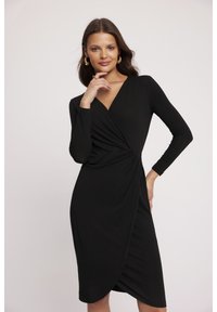 Czarna sukienka z długim rękawem, dekoltem w kształcie litery V i skręceniem w talii, wykonana z gładkiego, elastycznego materiału, sięgająca do kolan.