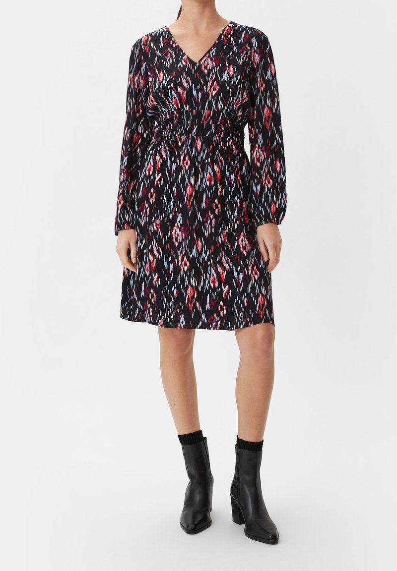 Femme portant une robe à motifs noire, rouge et blanche, à manches longues, longueur genou, avec des bottines noires, sur un fond uni.
