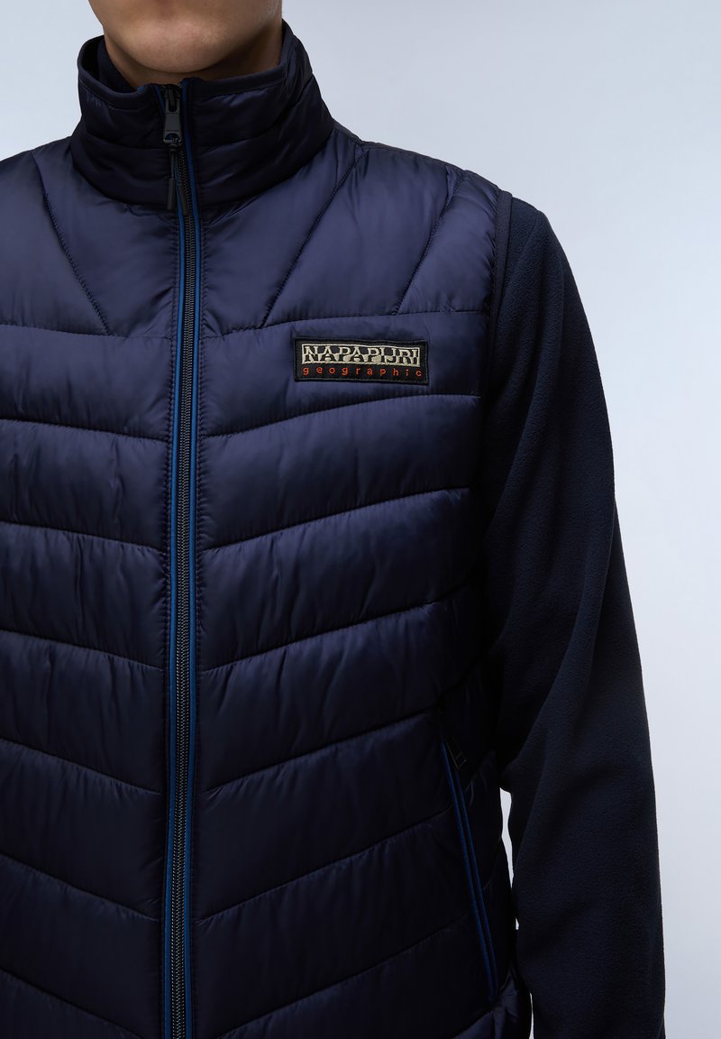 Gilet isolato blu navy con design trapuntato, colletto alto, zip frontale e patch con logo sul petto. Maniche e lati in pile.