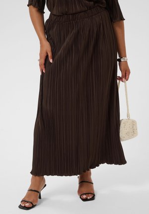 Femme portant une jupe longue plissée marron, des sandales à talons noires, un bracelet en argent et tenant un petit sac à main crème texturé.