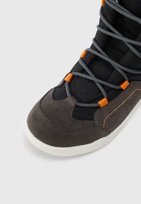 Zapatilla de deporte gris oscuro y negra con puntera gris y cordones grises pasando por ojales naranjas, y suela blanca sobre un fondo claro.