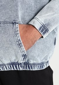Hellblaue Denim-Hoodie mit einer Kordelzug-Taille, elastischen Bündchen und einer Kängurutasche. Der Stoff hat ein ausgewaschenes Finish mit dunklen Akzenten.