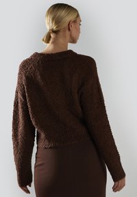 Kobieta z blond włosami w niskim kucyku, ubrana w teksturowany ciemno-brązowy sweter i pasującą spódnicę, odwrócona plecami przed gładkim tłem.