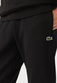 Svarta bomullsbyxor med en subtil Lacoste krokodil-logotyp i grönt och vitt, med en mjuk textur och elastisk midja.