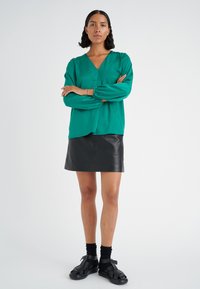 Blusa verde a maniche lunghe con scollatura a V e arricciature sulle maniche, abbinata a una mini gonna in pelle nera e sandali neri.