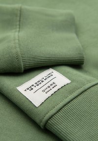 Tissu vert avec poignets côtelés et une étiquette blanche cousue affichant le texte : « VOTRE SEULE LIMITE EST VOTRE ESPRIT » et « OMBRE depuis 1985 ».