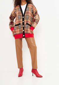 Cardigan de cuadros marrón con acentos rojos y ribete negro. Presenta grandes botones, bolsillos laterales y un corte relajado. Combinado con pantalones beige y tacones rojos.