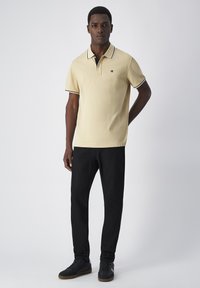 Lichtgeel polo shirt met zwarte kraag en mouwaccenten, gecombineerd met zwarte broek en zwarte sneakers, met een subtiele logo-detail.