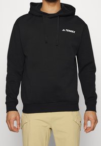Sudadera con capucha negra hecha de una mezcla de algodón. Cuenta con un bolsillo canguro en la parte frontal y un pequeño logo blanco "Terrex" en el pecho.