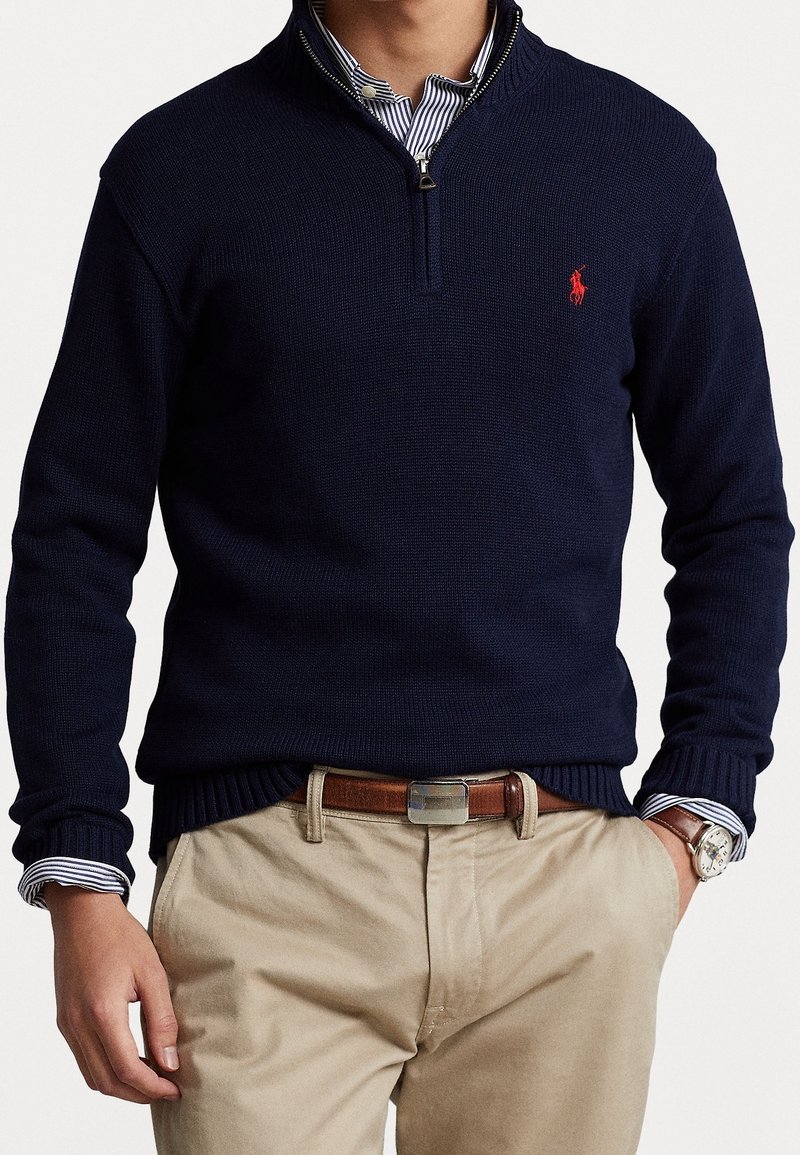 Homme portant un pull bleu marine à demi-fermeture éclair par-dessus une chemise rayée, pantalon beige, ceinture marron et montre argentée au poignet, mains dans les poches.
