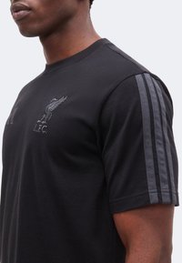 Άνδρας που φορεί μαύρο T-shirt της Liverpool FC με γκρι ρίγες στα μανίκια και το έμβλημα του συλλόγου στο στήθος, εμφανιζόμενος από τον ώμο μέχρι το μέσο του κορμού.