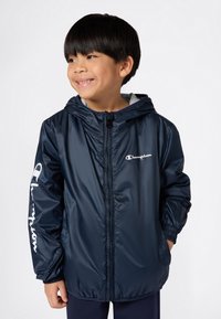 Junger Junge trägt eine marineblaue Champion Zip-Up Kapuzenjacke mit Logo auf der Brust und dem Ärmel, lächelt und schaut zur Seite.