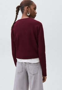 Burgundy stickad tröja med en kortad design, långa ärmar och rund halsringning, bärs över en vit topp, kombinerat med gråa jeans.