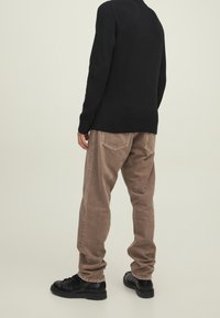 Pull noir côtelé, pantalon beige taille haute avec poches arrière, et chaussures noires à lacets. Vue de dos avec un fond neutre.