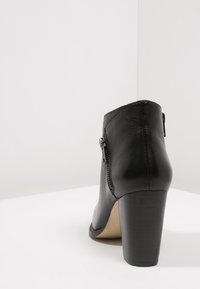 Zwarte leren enkelboot met zijrits en chunky hoge hak, gezien van achteren op een witte ondergrond.