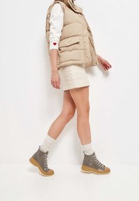 Beżowa pikowana kamizelka na białym swetrze z czerwonym sercem, w połączeniu z kremową spódnicą i szarymi sznurowanymi botkami na grubych beżowych podeszwach.