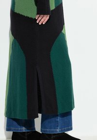Strickkleid mit geometrischen Mustern in Grün, Schwarz und Marineblau. Aushöhlungsschlitz am unteren Ende; kombiniert mit blauer Jeans und schwarzen Stiefeletten.