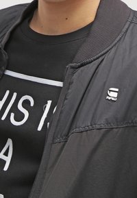 Czarna, lekka kurtka z fakturowanej tkaniny, z prążkowanym kołnierzem i małym logo na piersi, nałożona na czarną koszulkę z grafiką.