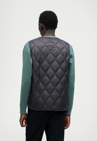 Δερματόπανο μαύρο γιλέκο με διαμαντένιο σχέδιο, που έχει ψηλό λαιμό και δύο κουμπιά με σνάπ στην κάτω πλευρά, φορεμένο πάνω από μία τιυαλέτα μπλούζα με μακριά μανίκια σε απόχρωση τιρκουάζ.