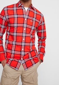 Camicia a quadri rossi con accenti blu e bianchi, dotata di una singola tasca sul petto e maniche lunghe, abbinata a pantaloni cargo kaki.