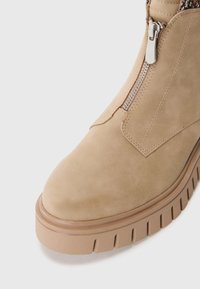 Beige suede ankle boot featuring a textured upper, side zipper, and a chunky, ridged rubber sole for grip and durability.