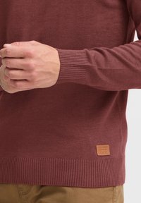 Pull à mailles bordeaux avec des poignets et un ourlet côtelés. Présente une petite étiquette en cuir en bas avec du texte. Le matériau semble lisse et doux.