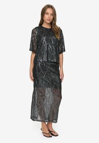 Kvinde iført en sort semi-gennemsigtig sequin-top og matchende lang nederdel med tynde sorte sandaler, stående mod en helt hvid baggrund.