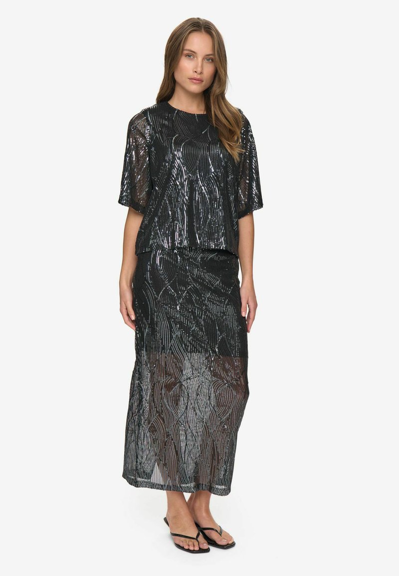 Kvinde iført en sort semi-gennemsigtig sequin-top og matchende lang nederdel med tynde sorte sandaler, stående mod en helt hvid baggrund.