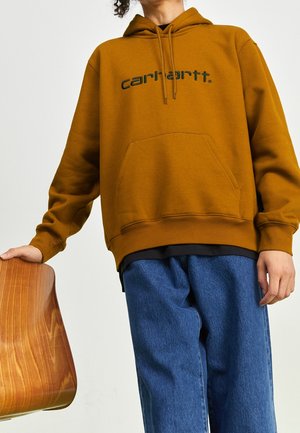 Person iført sennepsfarvet Carhartt-hoodie og blå jeans, hviler hånden på en træstol mod en ensfarvet lys baggrund.