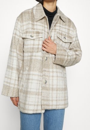 Beige und weiße karierte Wolljacke mit Knopfverschluss, Brusttaschen, getragen über einem schwarzen Rollkragenpullover und blauen Jeans.