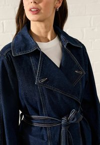 Denim kaput u tamnoplavoj boji s velikim ovratnikom, pojasom u struku i vidljivim šavovima; model nosi bijelu majicu ispod.