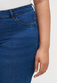 Blue denim jeans featuring a slim fit, detailed stitching, five-pocket design, and a medium-rise waistband with a button closure.