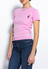 Różowa dopasowana koszulka z miękkiej bawełny, z małym czarnym logo na lewym piersi, połączona z ciemnoniebieskimi dżinsami.