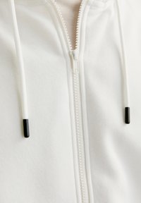 Hoodie zippé blanc en tissu texturé, avec une fermeture éclair centrale, des cordons de serrage avec des embouts noirs et un design minimaliste.