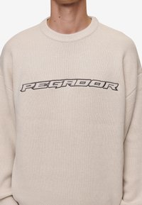 Beige stickad tröja med rund halsringning och broderad logotyp "PEGADOR" i mörkgrått över bröstet. Har ribbade manschetter och nederkant.
